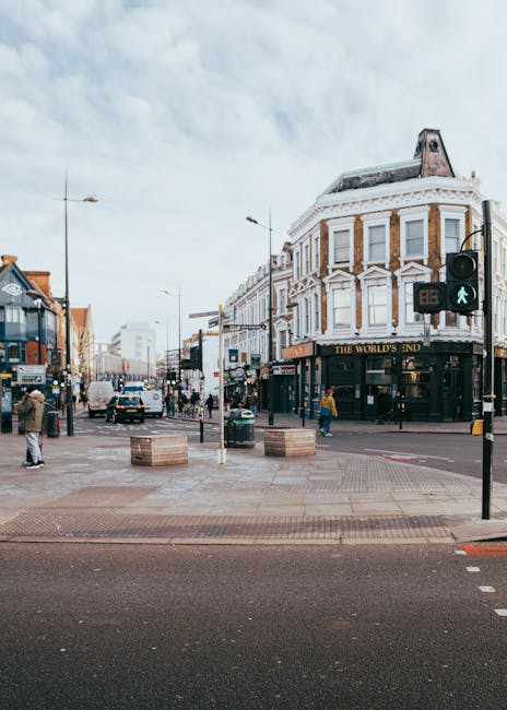 A busy urban street corner featuring a four-story building with ornate architectural details, including large bay windows with white trim and a rounded corner facade, housing a pub named 'The World's End' on the ground floor. The surrounding area includes pedestrians crossing the street, some waiting at a traffic light showing a green pedestrian signal. There are parked cars along the roadside, and the street is lined with modern and older style buildings. In the foreground, there are wooden planter boxes placed on the pavement, and streetlights and lampposts are visible, contributing to the lively city atmosphere with an overcast sky overhead. This setting may be typical for areas served by private waste removal services, such as Waste Clearance Camden, especially when managing rubbish collected from commercial or residential premises in the vicinity.