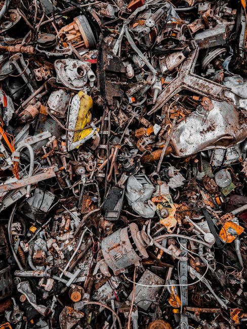 An extensive pile of various discarded automotive parts and metal debris, including engine blocks, belts, and smaller components, all showing signs of rust, corrosion, and dirt. The metal surfaces vary in texture, from smooth painted finishes to rough, corroded patches, with colors predominantly in shades of dark gray, brown, and rust-orange. The debris is densely packed and appears to be part of a scrap metal collection or waste clearance, situated outdoors on a gravel or dirt surface. Some wires and cables are tangled among the parts, and a few pieces have bright yellow or orange markings or labels. The lighting is natural, creating subtle shadows and highlights that accentuate the textures and disarray of the scrap metal. This scene is typical of a vehicle dismantling yard or a site where non-recyclable waste has been collected for transportation or disposal, illustrating a situation where private waste clearance services like Waste Clearance Camden may be involved in handling this type of bulky automotive rubbish.