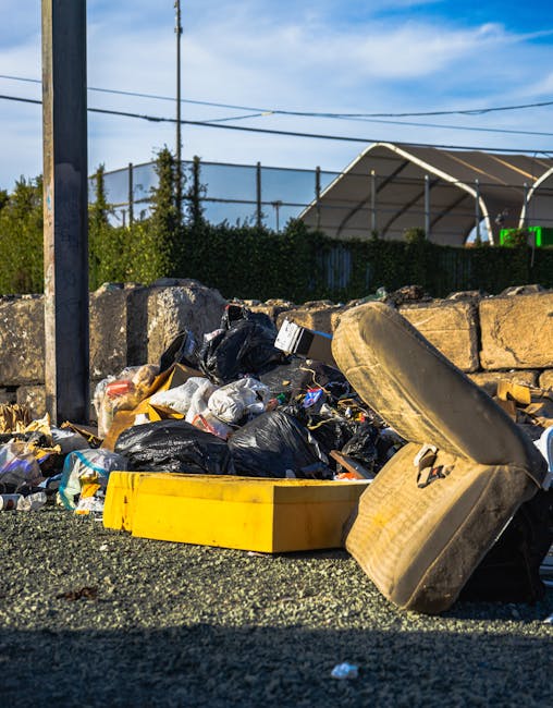An outdoor scene showing a pile of mixed household rubbish and waste materials accumulated on a gravel surface. The collection includes black plastic bin bags, a large yellow plastic container or toolbox, a worn beige or light brown car seat, and various smaller items such as discarded packaging and debris. The waste is scattered in front of a low stone wall made of large, roughly hewn blocks, which separates the area from a background featuring a fenced tennis court and some greenery, including trees and bushes. Overhead, there are power lines and a partly cloudy sky with patches of blue, suggesting a sunny day. The image captures the cluttered state of rubbish awaiting collection or removal, reflecting services offered by waste clearance companies like Waste Clearance Camden, including possible alternatives to council bulky waste disposal for private or onsite clearance.