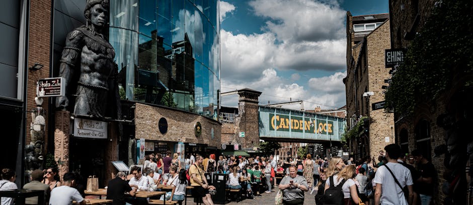 A busy urban scene at Camden Market featuring a diverse crowd of pedestrians seated at outdoor tables and standing along the pavement. In the foreground, several individuals are engaged in eating, drinking, or talking, with some wearing casual clothing and sunglasses. To the left, a large bronze statue of a figure in period attire stands mounted on a brick wall, adjacent to a modern glass-fronted building that reflects the sky and nearby structures. The background shows a weathered brick building partially shaded by trees, contrasting with the contemporary glass architecture. A prominent green sign reading 'CAMDEN LOCK' spans across the street, suspended above the crowd, indicating the market area. The overall environment is lively with a mix of natural daylight and overcast sky, highlighting the retail and social atmosphere typical of independent markets and informal waste handling, with some visible tables and elements suggestive of outdoor dining or gatherings, while Waste Clearance Camden's business focus remains aligned with the site's waste and rubbish removal context.