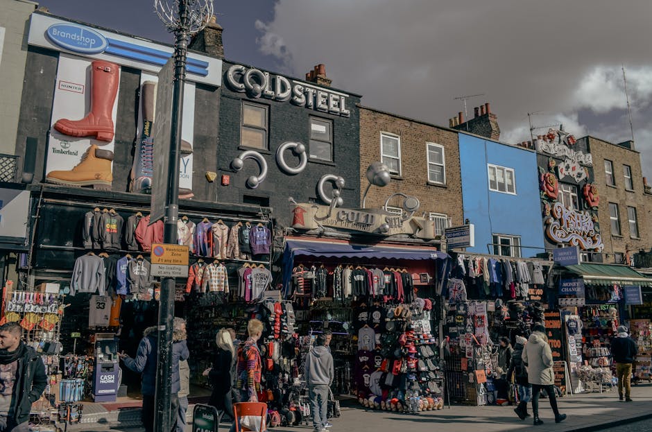 A busy urban scene at Camden Market featuring a diverse crowd of pedestrians seated at outdoor tables and standing along the pavement. In the foreground, several individuals are engaged in eating, drinking, or talking, with some wearing casual clothing and sunglasses. To the left, a large bronze statue of a figure in period attire stands mounted on a brick wall, adjacent to a modern glass-fronted building that reflects the sky and nearby structures. The background shows a weathered brick building partially shaded by trees, contrasting with the contemporary glass architecture. A prominent green sign reading 'CAMDEN LOCK' spans across the street, suspended above the crowd, indicating the market area. The overall environment is lively with a mix of natural daylight and overcast sky, highlighting the retail and social atmosphere typical of independent markets and informal waste handling, with some visible tables and elements suggestive of outdoor dining or gatherings, while Waste Clearance Camden's business focus remains aligned with the site's waste and rubbish removal context.