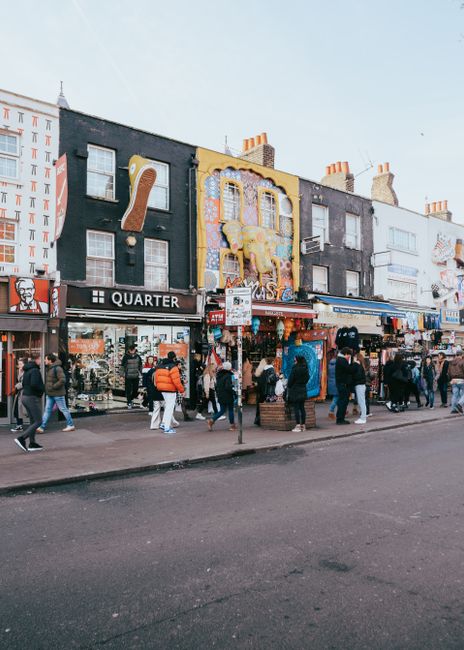 A busy street scene in Camden featuring a row of multi-storey shops with painted facades in various colors, including dark blue, gray, and a bright yellow building with decorative mural art. The ground level includes retail stores such as a clothing shop with signage, and there are several pedestrians walking along the pavement, some carrying shopping bags or wearing jackets. In front of the shops, a small stand displays goods, and a lamppost with street signs and posters is centrally positioned. The street surface appears asphalt with visible markings, and the overall atmosphere suggests an active commercial area. This setting illustrates a typical urban environment where private waste handling or alternative rubbish collection services, like those provided by Waste Clearance Camden, may be employed to manage waste generated by shop patrons and businesses, especially outside conventional municipal refuse collection times or locations.