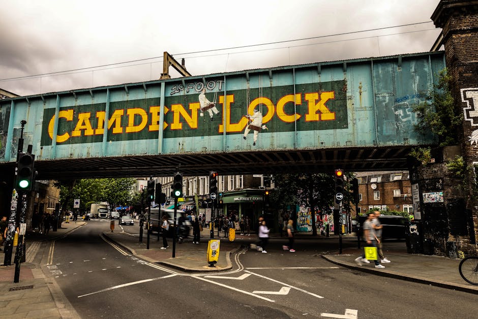 A large, weathered metal railway bridge painted in faded turquoise spans across a busy urban street, with the words 'Camden Lock' prominently displayed in bold yellow letters on its side. The sign includes a small illustration of two children hanging from the word 'Camden,' slightly peeling and showing signs of rust and age. The bridge is supported by brick pillars on either side, with graffiti and patches of climbing plants visible on the right side. Below, the street is filled with pedestrians crossing at traffic lights, some blurred from movement, and several vehicles including cars and bicycles, indicating a lively and active setting. The pavement is lined with black bollards and yellow plastic barriers, with shopfronts and graffiti-covered walls in the background. The scene is under overcast skies, creating a muted, diffuse lighting that highlights the textures of the metal and brick surfaces. This urban environment illustrates a typical scene where local waste management or rubbish removal services, such as Waste Clearance Camden, may be involved in maintaining the cleanliness of busy city areas adjacent to historic structures like this bridge, emphasizing the importance of private disposal and on-site clearance solutions.