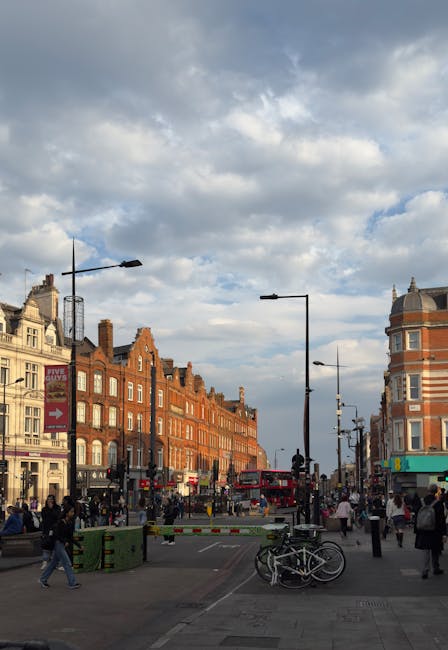 A busy street scene in Camden featuring a row of multi-storey shops with painted facades in various colors, including dark blue, gray, and a bright yellow building with decorative mural art. The ground level includes retail stores such as a clothing shop with signage, and there are several pedestrians walking along the pavement, some carrying shopping bags or wearing jackets. In front of the shops, a small stand displays goods, and a lamppost with street signs and posters is centrally positioned. The street surface appears asphalt with visible markings, and the overall atmosphere suggests an active commercial area. This setting illustrates a typical urban environment where private waste handling or alternative rubbish collection services, like those provided by Waste Clearance Camden, may be employed to manage waste generated by shop patrons and businesses, especially outside conventional municipal refuse collection times or locations.