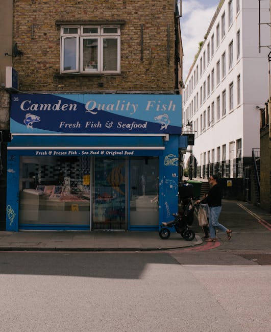 A storefront for Camden Quality Fish, a fresh fish and seafood shop, situated on a street corner with a blue sign displaying the business name and a fish graphic. The shop has large glass windows revealing refrigeration units inside and a metal door at the entrance. Above the shop, there is an upper-floor window with a white frame set into a brown brick building. To the right of the shop, a modern white multi-story building with large windows extends along the street. In front of the shop, a pedestrian is walking while pushing a stroller, dressed in a black top and light-colored trousers, with a beige bag hanging from the stroller handle. The sidewalk is made of concrete, and a curb separates it from the light gray tarmac road, which is empty of vehicles at this moment. The scene is illuminated by natural daylight, suggesting a clear weather condition. The overall setting indicates a typical urban street, potentially served by private waste collection services, relevant to local rubbish removal activities for nearby residents and businesses such as Waste Clearance Camden, particularly in the context of alternative waste handling methods like on-site clearance or independent collection.