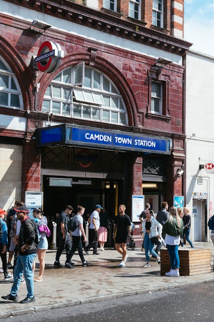 A storefront for Camden Quality Fish, a fresh fish and seafood shop, situated on a street corner with a blue sign displaying the business name and a fish graphic. The shop has large glass windows revealing refrigeration units inside and a metal door at the entrance. Above the shop, there is an upper-floor window with a white frame set into a brown brick building. To the right of the shop, a modern white multi-story building with large windows extends along the street. In front of the shop, a pedestrian is walking while pushing a stroller, dressed in a black top and light-colored trousers, with a beige bag hanging from the stroller handle. The sidewalk is made of concrete, and a curb separates it from the light gray tarmac road, which is empty of vehicles at this moment. The scene is illuminated by natural daylight, suggesting a clear weather condition. The overall setting indicates a typical urban street, potentially served by private waste collection services, relevant to local rubbish removal activities for nearby residents and businesses such as Waste Clearance Camden, particularly in the context of alternative waste handling methods like on-site clearance or independent collection.